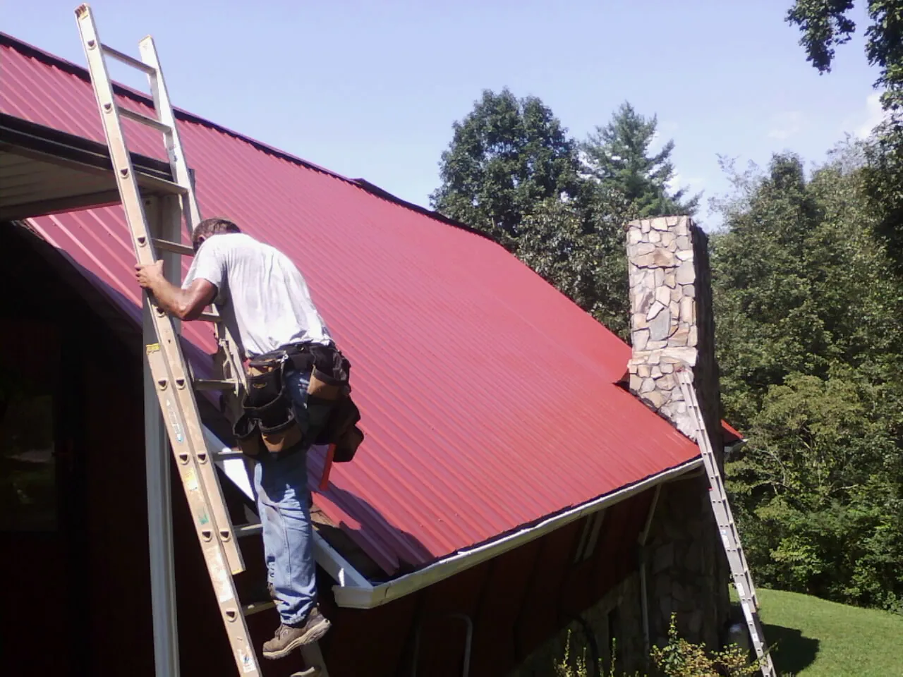 Roofer on a ladder beside a red metal roof and stone chimney — Soffit & Fascia Repair by Roofing Brothers in Howard Lake